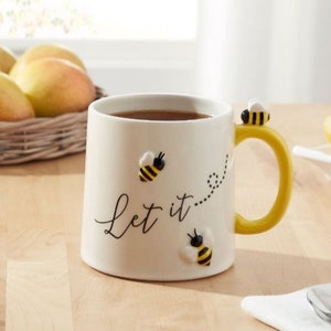 May include: A white ceramic mug with a yellow handle, featuring the black cursive text Let it, a dotted flight path, and three small, raised, three-dimensional bumblebee figures in yellow and black, with one bee perched on the top of the handle.