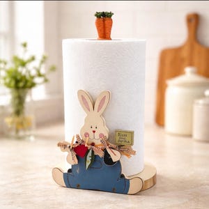 May include: A decorative paper towel holder featuring a wooden bunny design. The bunny wears blue overalls and holds a sign that reads "Bless our Garden." A carrot tops the paper towel roll. The holder sits on a wooden base.