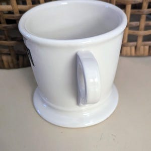 K&ouml;nnte beinhalten: Eine wei&szlig;e Rasierbecher aus Keramik mit Henkel und breitem Fu&szlig;. Der Becher hat ein schlichtes, klassisches Design mit leicht ausgestelltem Rand. Der Hintergrund zeigt einen Weidenkorb.