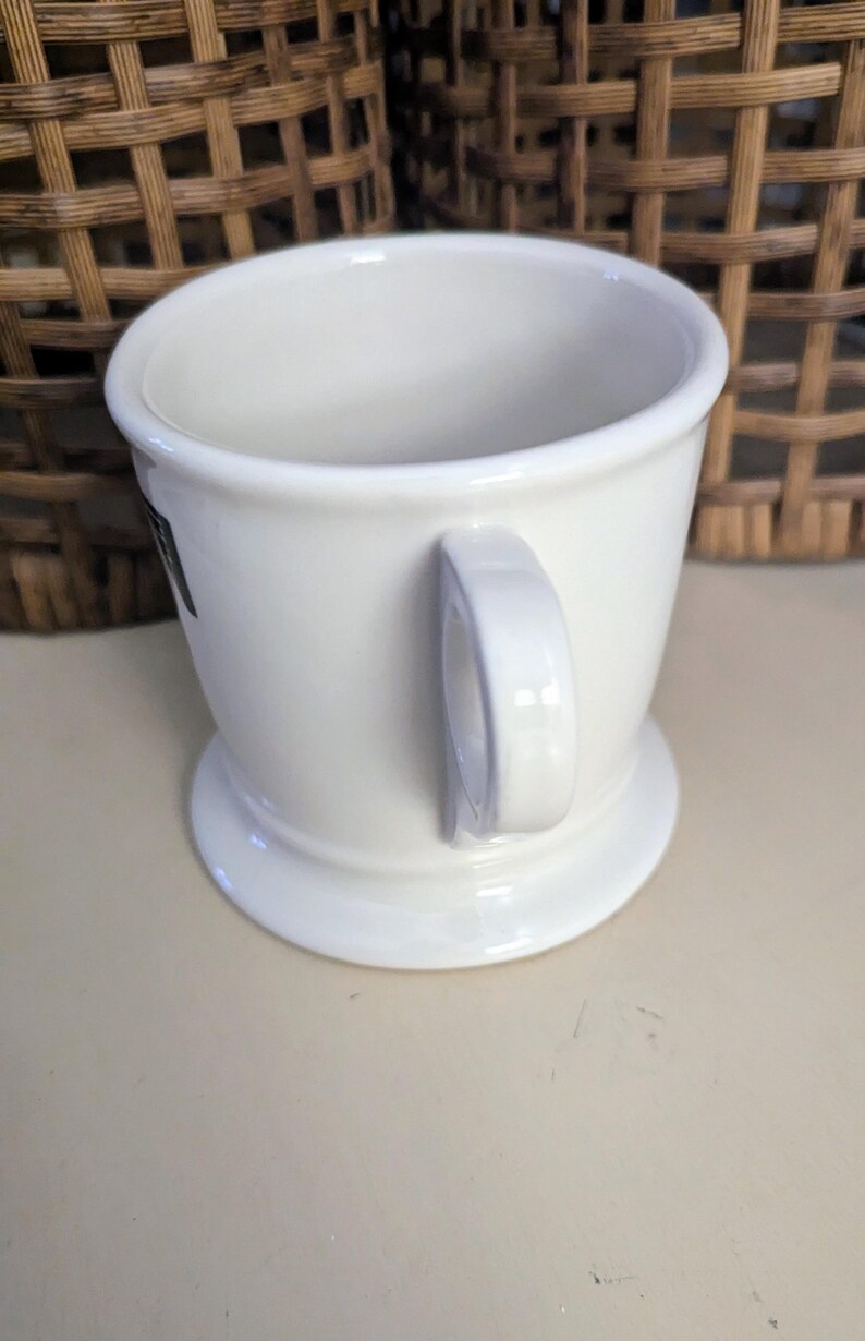 K&ouml;nnte beinhalten: Eine wei&szlig;e Rasierbecher aus Keramik mit Henkel und breitem Fu&szlig;. Der Becher hat ein schlichtes, klassisches Design mit leicht ausgestelltem Rand. Der Hintergrund zeigt einen Weidenkorb.