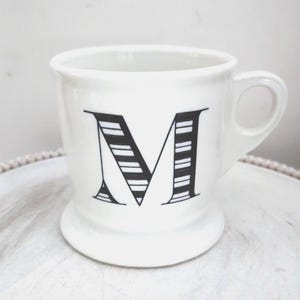 May include: White ceramic mug with a black letter "M" design. The letter is stylized with horizontal stripes. The mug has a handle and a pedestal base. The mug is on a white surface.