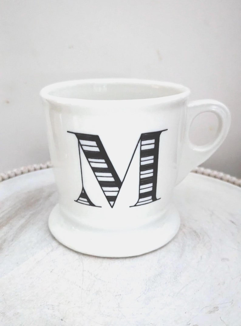 May include: White ceramic mug with a black letter "M" design. The letter is stylized with horizontal stripes. The mug has a handle and a pedestal base. The mug is on a white surface.