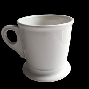 K&ouml;nnte beinhalten: Eine wei&szlig;e Keramiktasse mit rundem Boden und gebogenem Henkel. Die Tasse hat eine gl&auml;nzende Oberfl&auml;che und ein schlichtes, klassisches Design. Geeignet f&uuml;r Kaffee, Tee oder andere Getr&auml;nke.