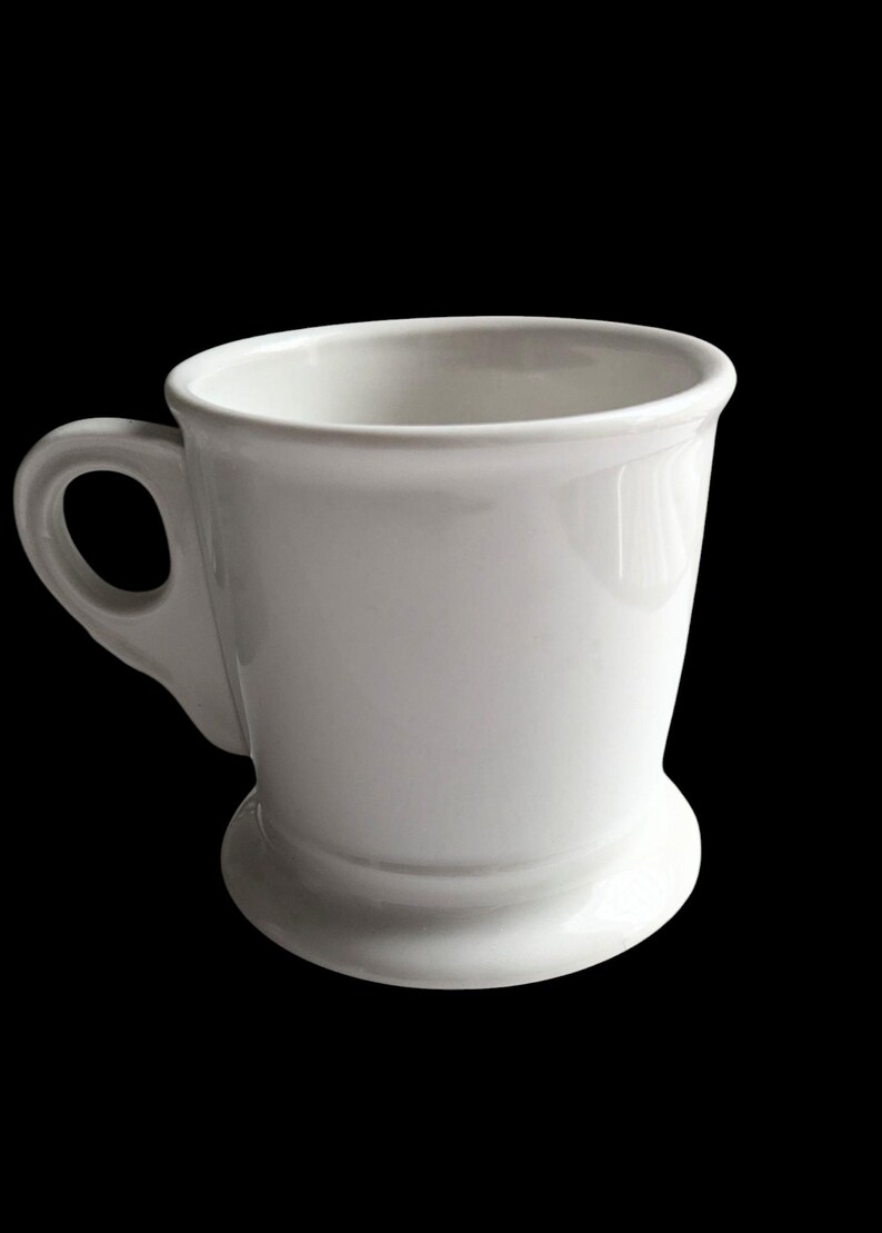 K&ouml;nnte beinhalten: Eine wei&szlig;e Keramiktasse mit rundem Boden und gebogenem Henkel. Die Tasse hat eine gl&auml;nzende Oberfl&auml;che und ein schlichtes, klassisches Design. Geeignet f&uuml;r Kaffee, Tee oder andere Getr&auml;nke.