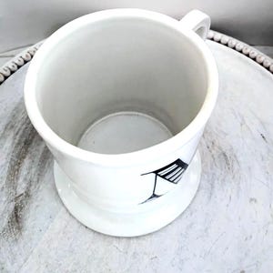 May include: White ceramic shaving mug with a handle and a wide base. The mug features a black and white graphic design on the side. The mug is empty and sits on a white surface.