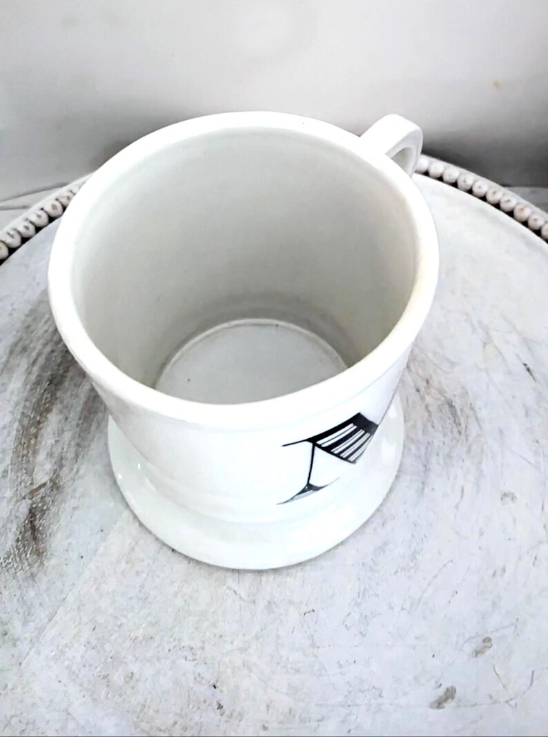 May include: White ceramic shaving mug with a handle and a wide base. The mug features a black and white graphic design on the side. The mug is empty and sits on a white surface.
