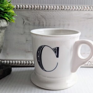 K&ouml;nnte beinhalten: Wei&szlig;e Keramiktasse mit gro&szlig;em schwarzem Buchstaben "C" auf der Vorderseite. Die Tasse hat einen abgerundeten Boden und einen Henkel. Im Hintergrund eine kleine gr&uuml;ne Pflanze in einem grauen Topf und ein wei&szlig;er Zierrahmen.