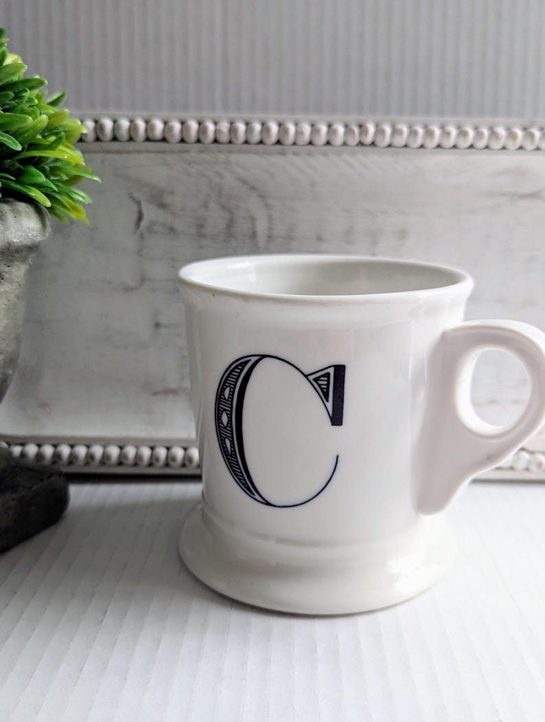 K&ouml;nnte beinhalten: Wei&szlig;e Keramiktasse mit gro&szlig;em schwarzem Buchstaben "C" auf der Vorderseite. Die Tasse hat einen abgerundeten Boden und einen Henkel. Im Hintergrund eine kleine gr&uuml;ne Pflanze in einem grauen Topf und ein wei&szlig;er Zierrahmen.