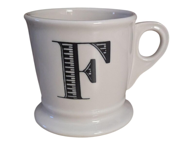 K&ouml;nnte beinhalten: Eine wei&szlig;e Keramiktasse mit dem schwarzen Buchstaben "F" auf der Vorderseite. Die Tasse hat einen abgerundeten Boden, einen gebogenen Henkel und einen leicht ausgestellten Rand. Der Buchstabe "F" ist mit vertikalen Linien gestaltet.