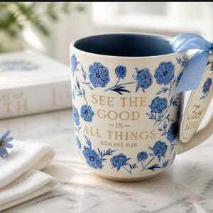May include: Cream-colored ceramic mug with blue floral design and the text "SEE THE GOOD IN ALL THINGS" from Romans 8:28. A light blue ribbon and tag are attached to the handle. A book and flowers are in the background.