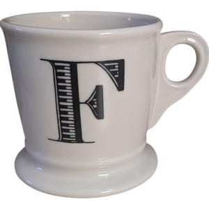 K&ouml;nnte beinhalten: Wei&szlig;er Keramikbecher mit einem schwarzen Buchstaben "F" auf der Vorderseite. Der Becher hat einen abgerundeten Boden, einen Henkel und einen leicht ausgestellten Rand. Der Buchstabe "F" ist mit vertikalen Linien und einem horizontalen Akzent gestaltet.