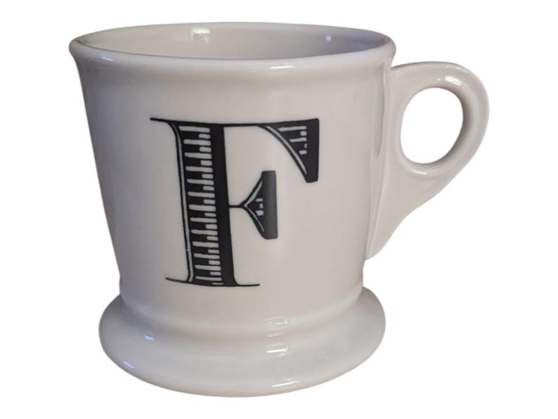 K&ouml;nnte beinhalten: Wei&szlig;er Keramikbecher mit einem schwarzen Buchstaben "F" auf der Vorderseite. Der Becher hat einen abgerundeten Boden, einen Henkel und einen leicht ausgestellten Rand. Der Buchstabe "F" ist mit vertikalen Linien und einem horizontalen Akzent gestaltet.