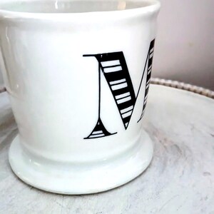 May include: White ceramic mug with a wide base and a large black letter "M" on the side. The letter is stylized with horizontal lines. The mug has a simple, elegant design.