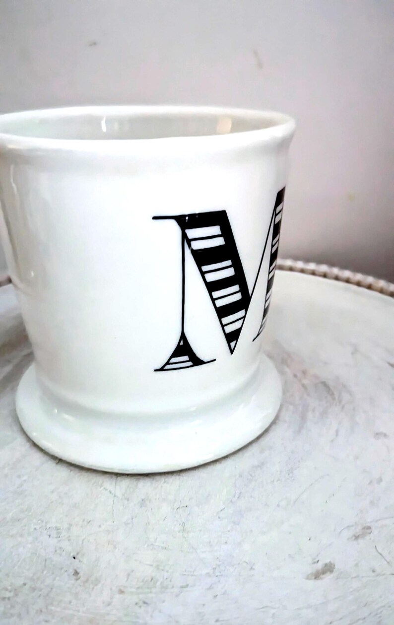 May include: White ceramic mug with a wide base and a large black letter "M" on the side. The letter is stylized with horizontal lines. The mug has a simple, elegant design.