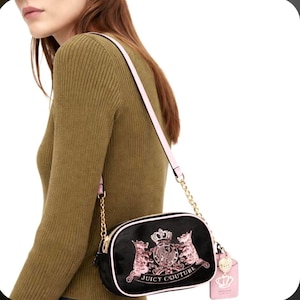 May include: Black crossbody bag with a pink strap and gold chain detail. The bag features a pink and gold crest design with the words "Juicy Couture". A pink tag with a crown charm hangs from the bag.