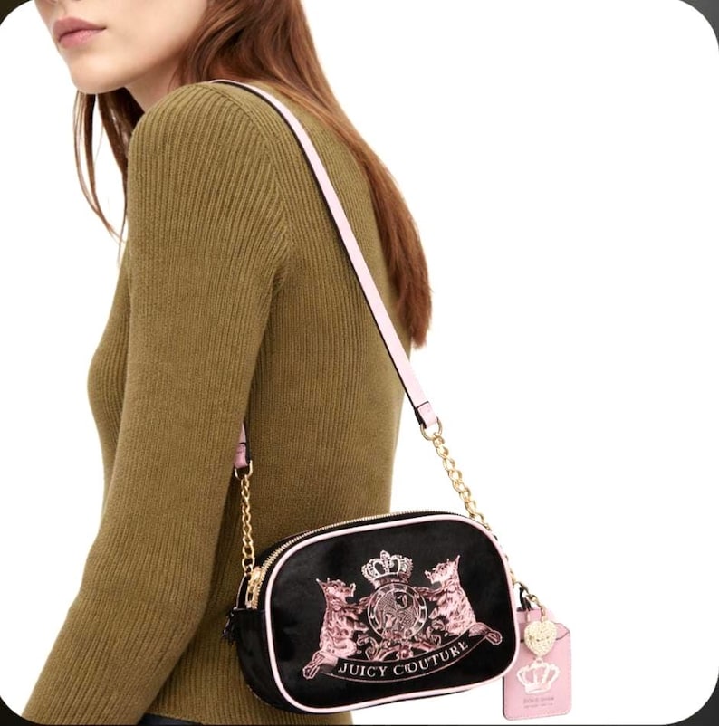 May include: Black crossbody bag with a pink strap and gold chain detail. The bag features a pink and gold crest design with the words "Juicy Couture". A pink tag with a crown charm hangs from the bag.