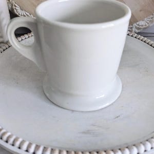 K&ouml;nnte beinhalten: Eine wei&szlig;e Kaffeetasse aus Keramik mit Henkel, die auf einem wei&szlig;en, runden Dekotablett mit Perlenrand steht. Die Tasse hat ein klassisches Design und einen breiten Boden. Das Tablett steht auf einer hellen Holzoberfl&auml;che.