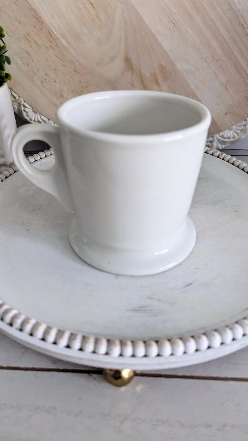 K&ouml;nnte beinhalten: Eine wei&szlig;e Kaffeetasse aus Keramik mit Henkel, die auf einem wei&szlig;en, runden Dekotablett mit Perlenrand steht. Die Tasse hat ein klassisches Design und einen breiten Boden. Das Tablett steht auf einer hellen Holzoberfl&auml;che.
