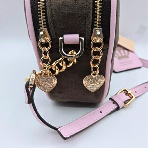 May include: A small, brown velour crossbody bag with a pink trim and strap. The bag features gold-tone zippers, chain details, and heart-shaped charms. The strap has a gold buckle. The bag has the text "Juicy Couture" on the heart charms.