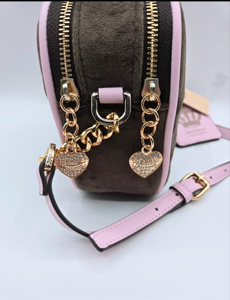 May include: A small, brown velour crossbody bag with a pink trim and strap. The bag features gold-tone zippers, chain details, and heart-shaped charms. The strap has a gold buckle. The bag has the text "Juicy Couture" on the heart charms.