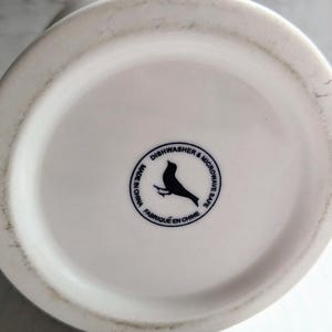 K&ouml;nnte beinhalten: Boden einer wei&szlig;en Keramiktasse mit einem schwarzen Vogellogo und dem Text "Sp&uuml;lmaschinen- und Mikrowellengeeignet" und "Made in China" in einem kreisf&ouml;rmigen Design.