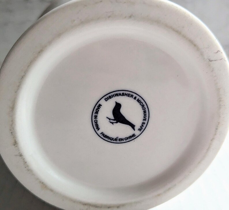 K&ouml;nnte beinhalten: Boden einer wei&szlig;en Keramiktasse mit einem schwarzen Vogellogo und dem Text "Sp&uuml;lmaschinen- und Mikrowellengeeignet" und "Made in China" in einem kreisf&ouml;rmigen Design.