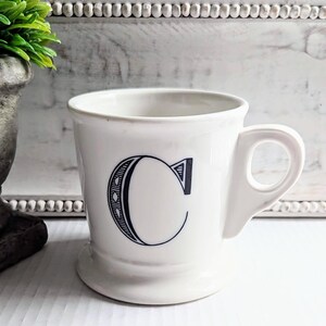 K&ouml;nnte beinhalten: Wei&szlig;e Keramiktasse mit einem gro&szlig;en, stilisierten schwarzen Buchstaben "C" auf der Vorderseite. Die Tasse hat einen abgerundeten Boden und einen Henkel. Der Hintergrund umfasst eine dekorative wei&szlig;e Holztafel und eine gr&uuml;ne Pflanze in einem grauen Topf.