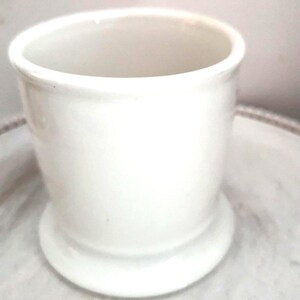 May include: A small, white ceramic container with a cylindrical body and a flared base. The container has a smooth, glossy finish and is set on a light-colored surface. The design is simple and classic, suitable for various uses.