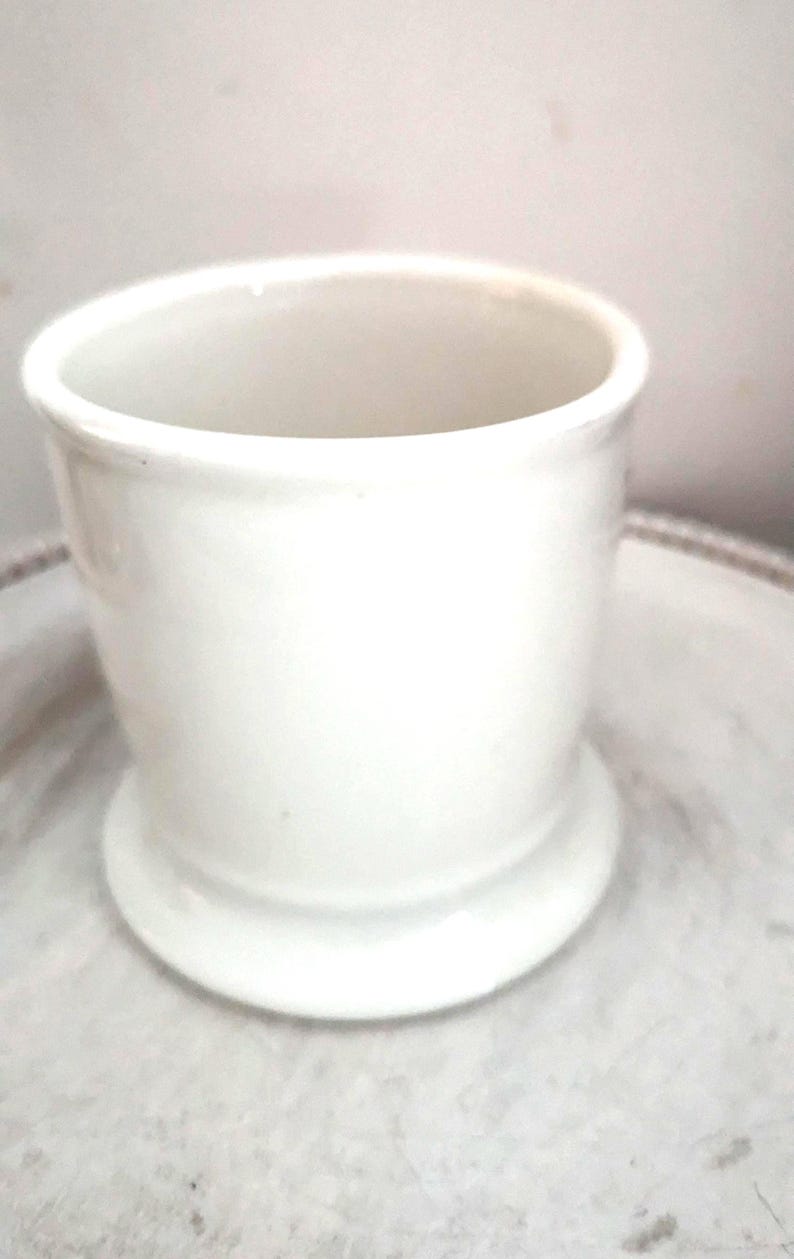 May include: A small, white ceramic container with a cylindrical body and a flared base. The container has a smooth, glossy finish and is set on a light-colored surface. The design is simple and classic, suitable for various uses.
