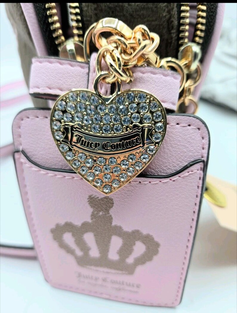 May include: A pink, small purse with a gold-colored chain and zipper. A heart-shaped charm with the words "Juicy Couture" is attached. The charm is encrusted with clear rhinestones. A crown design is visible on the front of the purse.