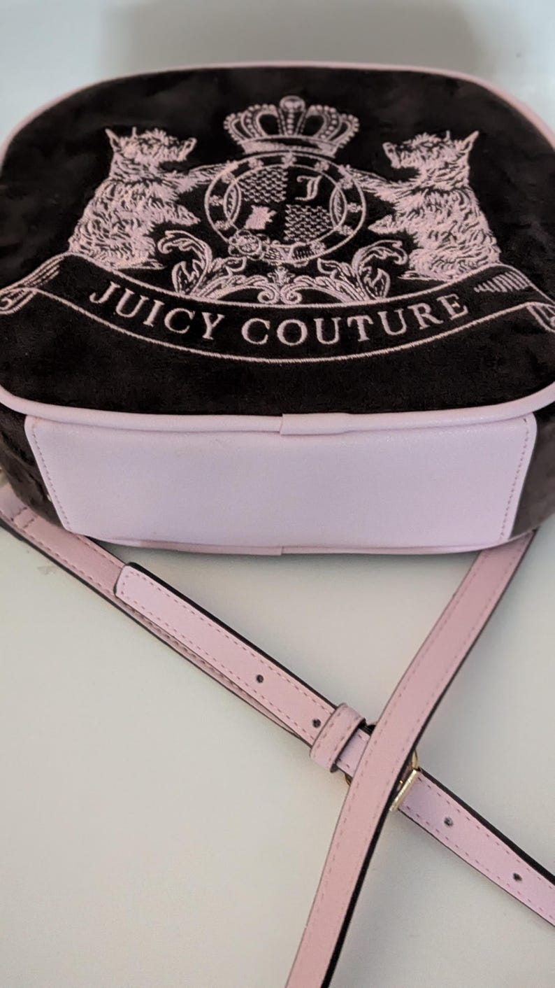 May include: A black and pink Juicy Couture crossbody bag. The bag features a pink and black design with the words "Juicy Couture" and a crest with two pink lions. The bag has a pink strap and a light pink bottom.