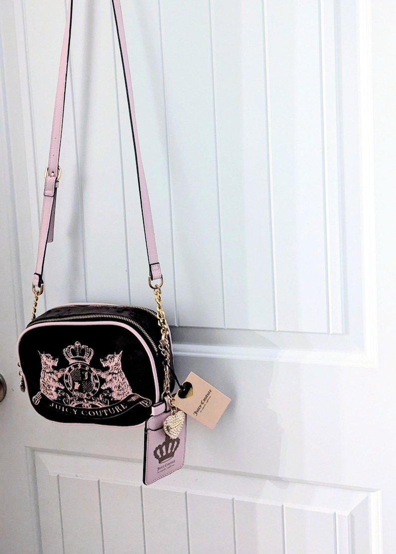 May include: Black crossbody bag with pink accents and gold-tone chain straps. The bag features a pink and gold emblem with the words "Juicy Couture". A matching pink cardholder and tag are attached.