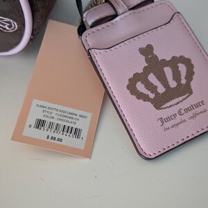 May include: A pink Juicy Couture cardholder with a gold-colored chain and bow detail. The cardholder features a crown design and the brand's logo. A tag with product information is also visible.
