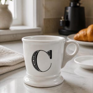 K&ouml;nnte beinhalten: Wei&szlig;e Keramik-Tasse mit einem schwarzen Buchstaben "C"-Design. Die Tasse hat einen abgerundeten Boden und einen gebogenen Henkel. Ein Croissant und eine Kaffeem&uuml;hle sind im Hintergrund. Die Tasse steht auf einer Marmorarbeitsplatte.