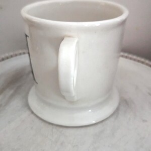 May include: A white ceramic shaving mug with a handle. The mug has a wide base and a slightly flared rim. The handle is a simple, curved design. The mug is clean and appears unused. The mug is likely for shaving.