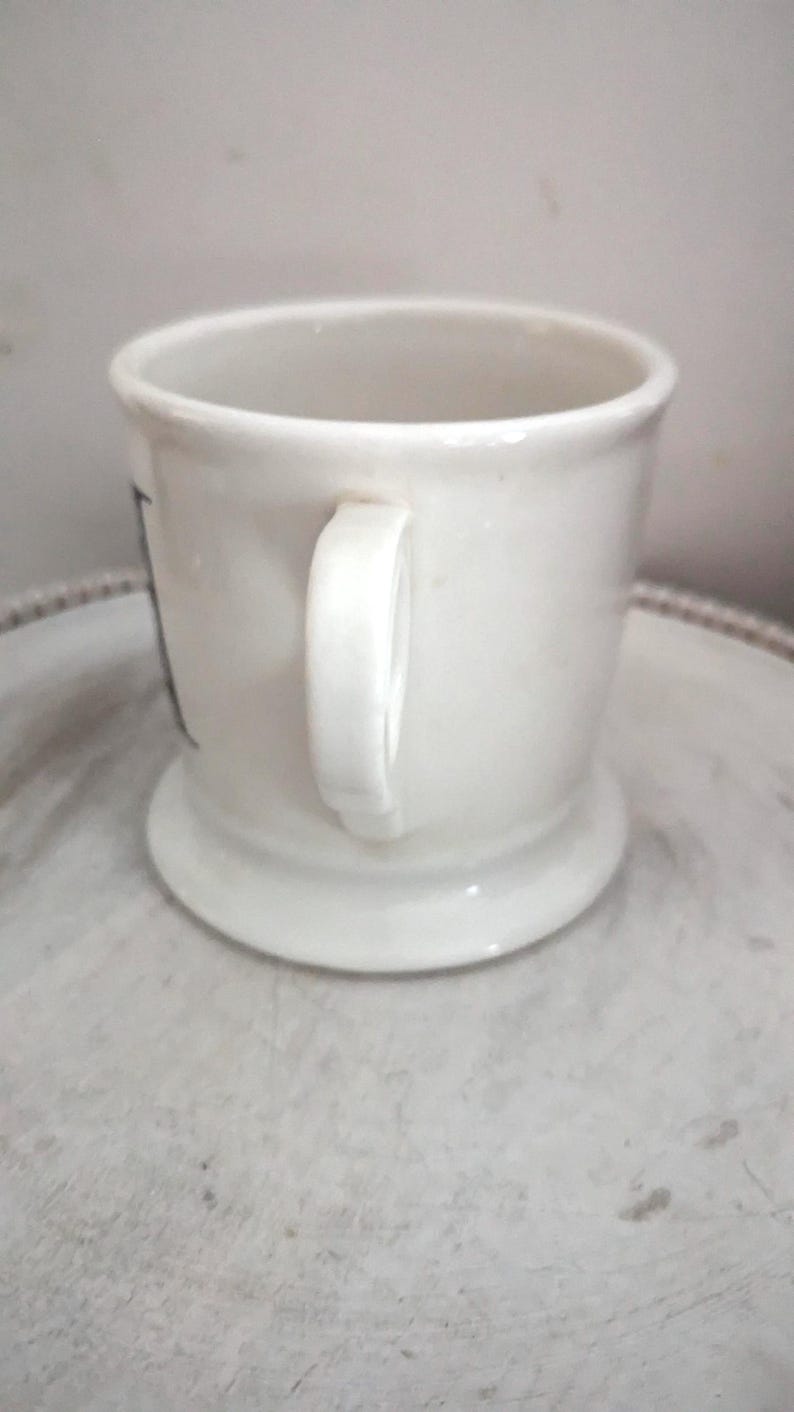 May include: A white ceramic shaving mug with a handle. The mug has a wide base and a slightly flared rim. The handle is a simple, curved design. The mug is clean and appears unused. The mug is likely for shaving.