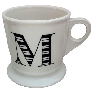 May include: White ceramic mug with a black and white striped letter "M" on the front. The mug has a rounded base, a curved handle, and a simple, elegant design. Perfect for coffee, tea, or any beverage.
