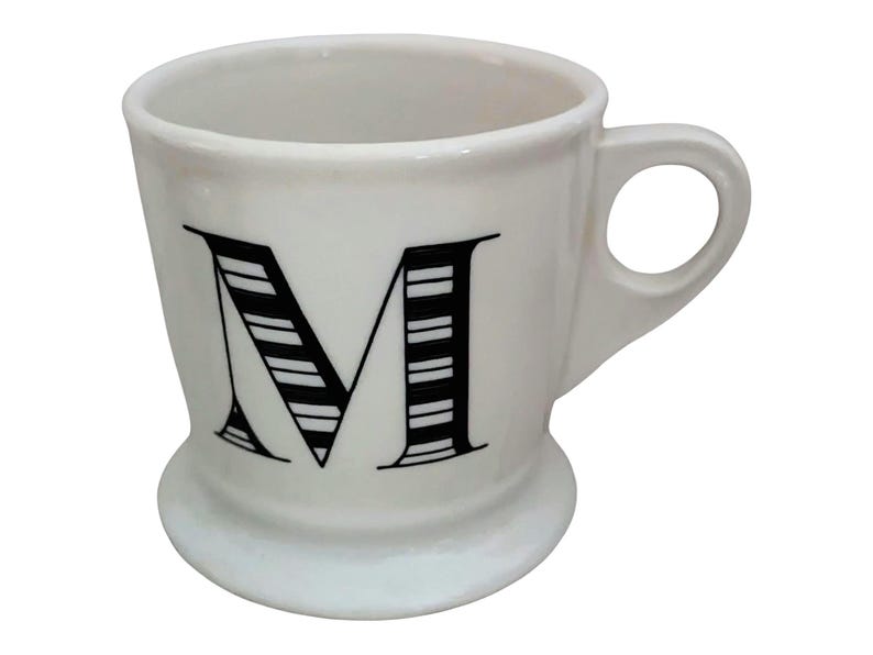 May include: White ceramic mug with a black and white striped letter "M" on the front. The mug has a rounded base, a curved handle, and a simple, elegant design. Perfect for coffee, tea, or any beverage.