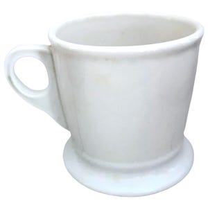 May include: A white ceramic shaving mug with a handle and a wide, circular base. The mug has a simple, classic design, perfect for holding shaving cream or soap. The handle is a loop shape, and the mug is clean and bright.