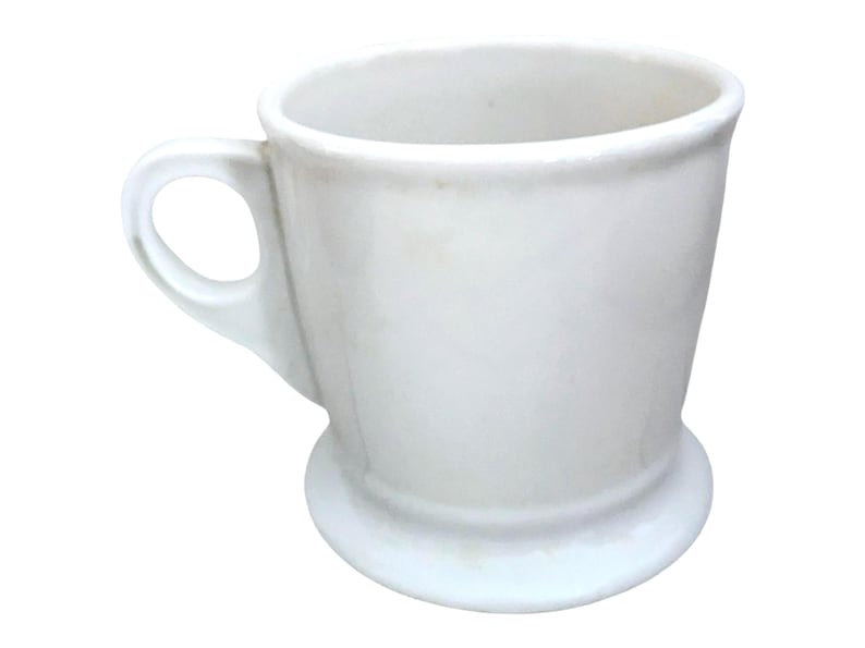 May include: A white ceramic shaving mug with a handle and a wide, circular base. The mug has a simple, classic design, perfect for holding shaving cream or soap. The handle is a loop shape, and the mug is clean and bright.