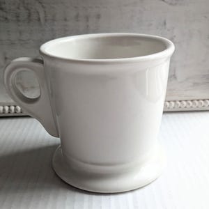 K&ouml;nnte beinhalten: Eine wei&szlig;e Keramiktasse mit rundem Henkel und Sockel. Die Tasse hat eine glatte, gl&auml;nzende Oberfl&auml;che und ein schlichtes, klassisches Design. Die Tasse eignet sich f&uuml;r Kaffee, Tee oder andere Getr&auml;nke.