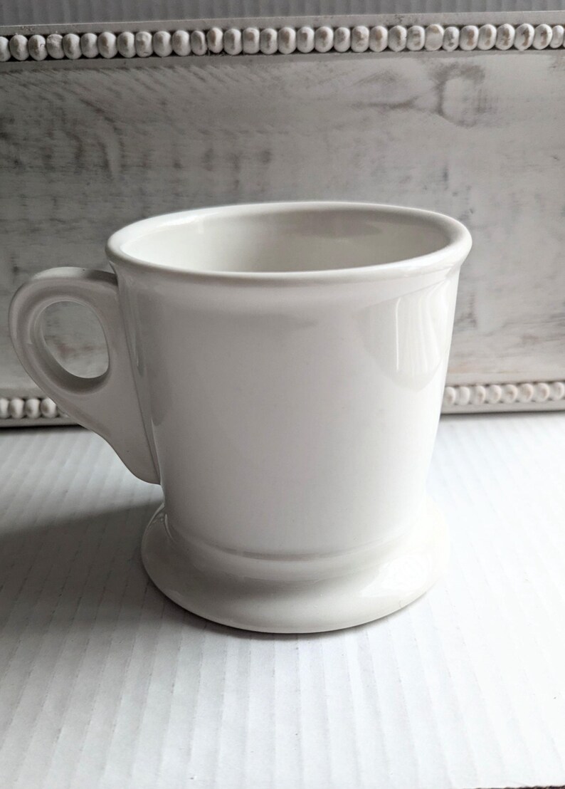 K&ouml;nnte beinhalten: Eine wei&szlig;e Keramiktasse mit rundem Henkel und Sockel. Die Tasse hat eine glatte, gl&auml;nzende Oberfl&auml;che und ein schlichtes, klassisches Design. Die Tasse eignet sich f&uuml;r Kaffee, Tee oder andere Getr&auml;nke.