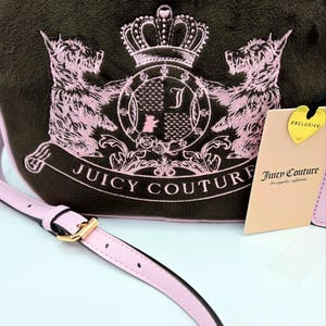 May include: A brown crossbody bag with a pink strap and trim. The bag features a pink embroidered design of two dogs, a crown, and the words "Juicy Couture". A tag reads "Exclusive".