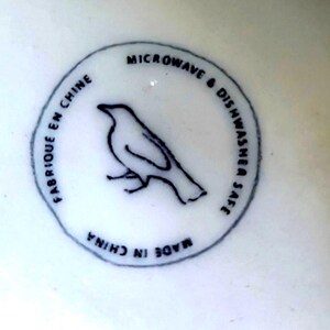 May include: A white ceramic surface with a circular black and white design. The design features a bird silhouette and the text "MICROWAVE & DISHWASHER SAFE", "FABRIQUE EN CHINE", and "MADE IN CHINA".