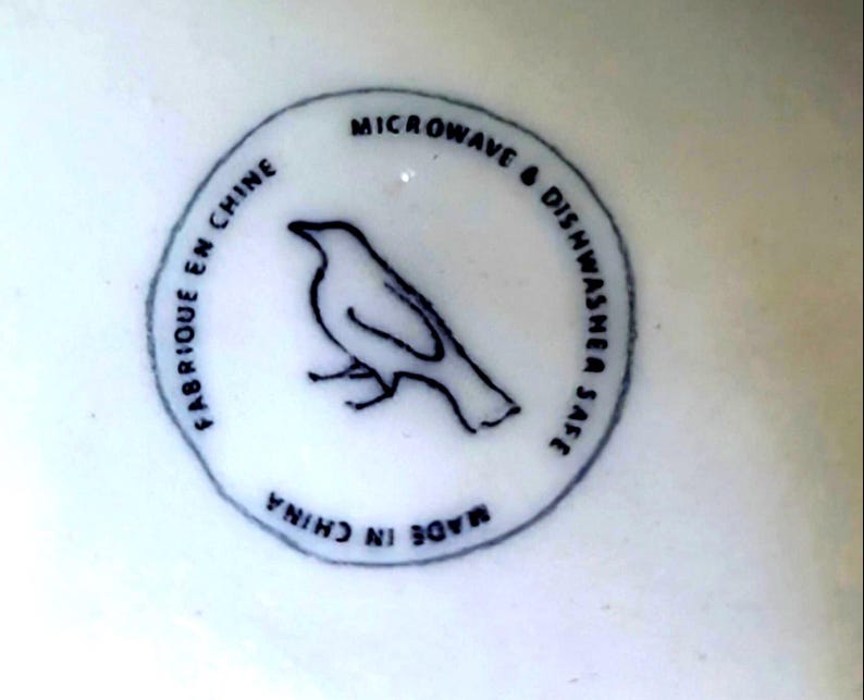 May include: A white ceramic surface with a circular black and white design. The design features a bird silhouette and the text "MICROWAVE & DISHWASHER SAFE", "FABRIQUE EN CHINE", and "MADE IN CHINA".