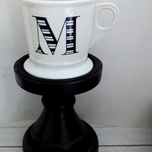 May include: A white ceramic mug with a black letter "M" design, set on a black decorative stand. The mug has a handle and a simple, elegant design. The stand is approximately 6 inches tall.