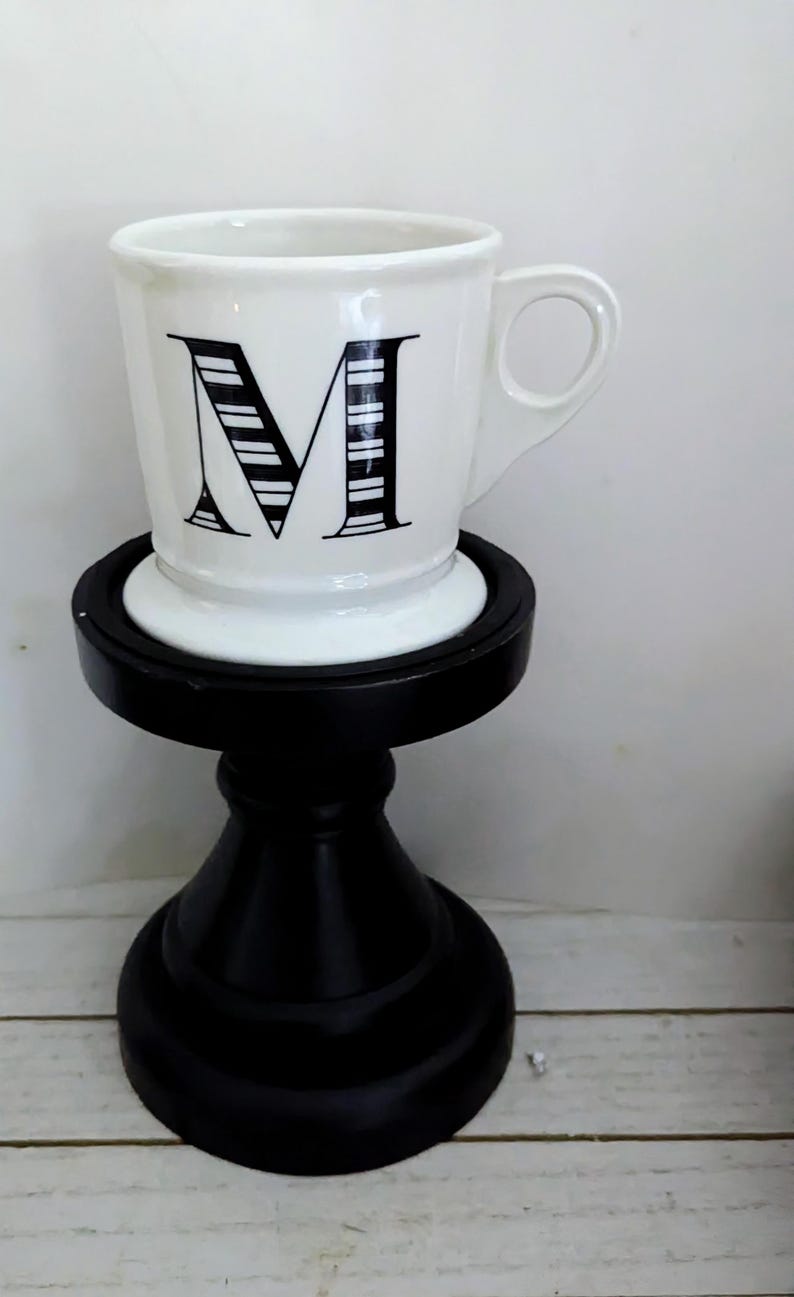 May include: A white ceramic mug with a black letter "M" design, set on a black decorative stand. The mug has a handle and a simple, elegant design. The stand is approximately 6 inches tall.