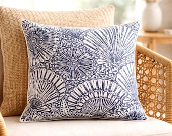 May include: A decorative pillow with a navy blue and white floral pattern. The pillow is square and features a detailed design of stylized flowers and fan-like shapes. It rests on a neutral-colored chair.