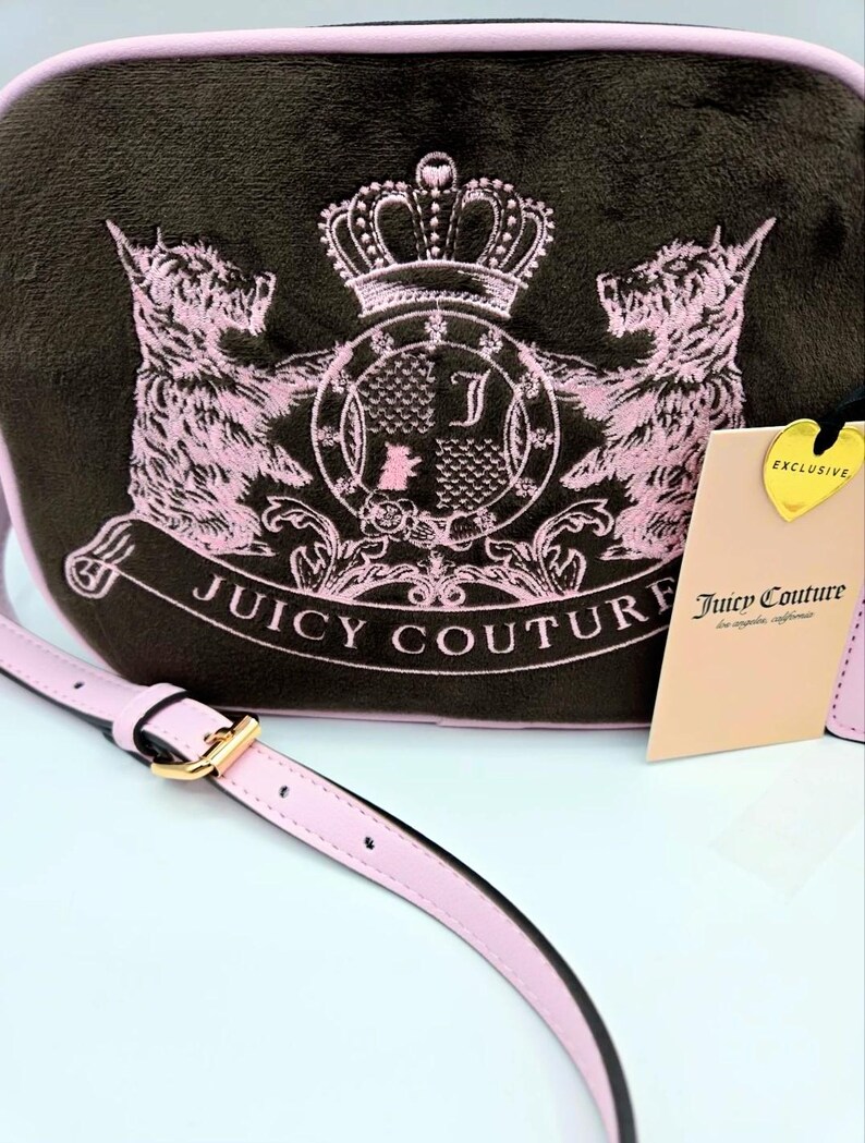 May include: A brown crossbody bag with a pink strap and trim. The bag features a pink embroidered design of two dogs, a crown, and the words "Juicy Couture". A tag with the words "Exclusive" and "Juicy Couture" is attached.