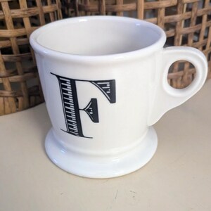 K&ouml;nnte beinhalten: Wei&szlig;e Keramiktasse mit schwarzem Buchstaben "F" auf der Vorderseite. Die Tasse hat einen Henkel und einen Sockel. Im Hintergrund ein Weidenkorb.